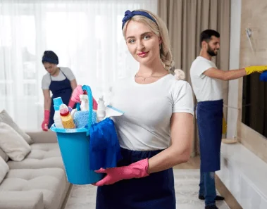Woman wearing gloves while holding a bucket full of cleaning supplies. In the background, there are two professional Airbnb cleaners dusting the living room.