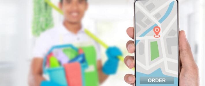 How to Find an Airbnb Cleaning Service in Your Area Person holding up a phone with a map. In the background there is a man with Airbnb cleaning products such as a mop and several cleaning agents.